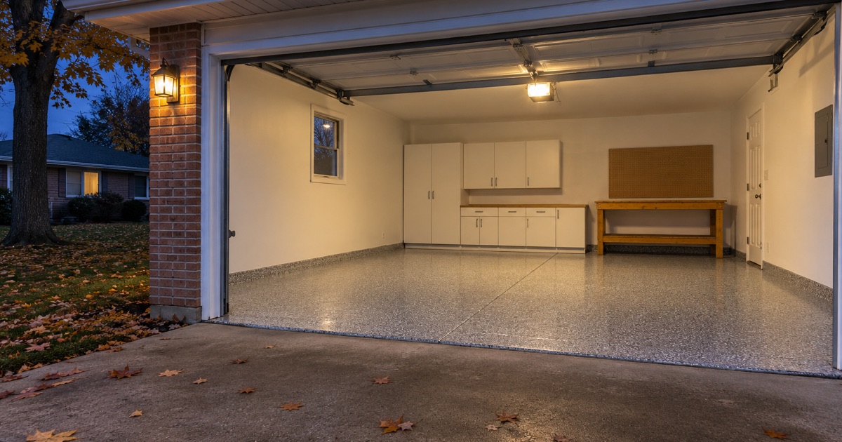 A finished gray flake floor in the open attached garage of a 1960s Waterloo ranch house at dusk, the overhead light on inside.